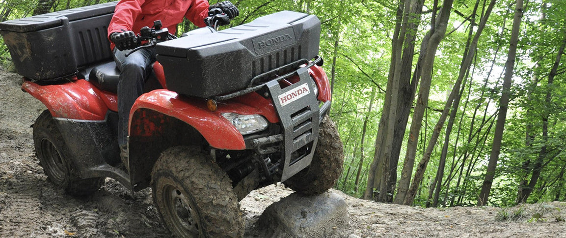 Honda TRX420FA6 all-terrain vehicle riding through a rugged trail, showcasing durable off-road design and Honda branding.
